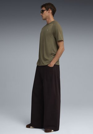 Mężczyzna w okularach przeciwsłonecznych, oliwkowozielonej koszulce z krótkim rękawem i szerokich czarnych spodniach, stojący bokiem z rękami w kieszeniach na jednolitym szarym tle.