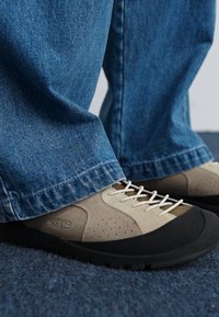 Chaussures d'escalade beiges avec des semelles en caoutchouc noires et un design perforé, associées à un pantalon en denim large. Les chaussures disposent de lacets blancs et d'un logo.