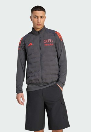 Uomo che indossa un gilet imbottito grigio scuro con i loghi rossi di Audi e Revolut sopra una camicia a maniche lunghe e pantaloncini cargo neri, in piedi con una mano in tasca.