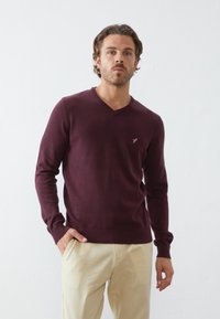 Burgundy V-hals trui gemaakt van zacht breimateriaal, met lange mouwen en een geribbelde zoom, voorzien van een klein logo op de linkerborst.