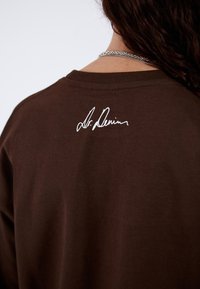 Camiseta de algodón marrón con cuello redondo y un estampado blanco de firma en la parte superior de la espalda. La tela parece suave con una textura blanda.