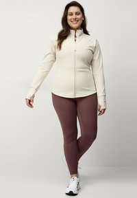 Crèmekleurige zip-up jack met lange mouwen, gecombineerd met donker bordeauxrode legging en witte sportschoenen; gladde textuur, aansluitend ontwerp.