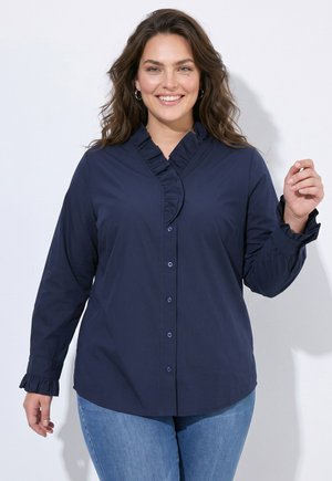Lächelnde Frau mit welligem braunem Haar, die eine dunkelblaue Bluse mit Knöpfen, Rüschenkragen und Manschetten sowie eine hellblaue Jeans trägt.