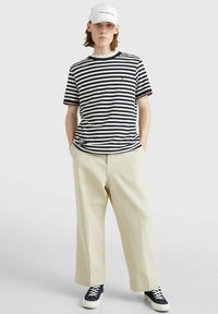 T-shirt a righe blu navy e crema con accenti rossi, pantaloni larghi crema e sneakers blu navy. Un cappellino bianco completa l'outfit.