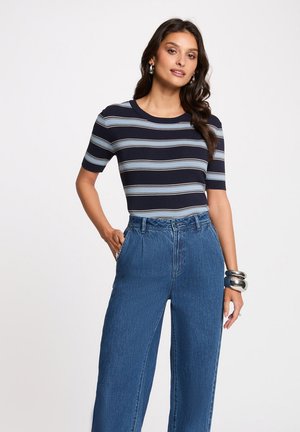 Frau mit langen, welligen, dunklen Haaren, die silberne Creolen, ein gestreiftes marine- und hellblaues Kurzarmshirt, hoch taillierte blaue Jeans und silberne Armbänder trägt.