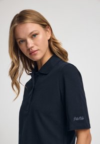 Granatowa koszulka polo z kołnierzykiem i krótkimi rękawami, wykonana z teksturowanej tkaniny, z zapięciem na trzy guziki. Na rękawie haft "Polo Club".