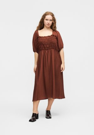 Γυναίκα με μακριά σγουρά μαλλιά, φοράει καφέ μίντι φόρεμα με πιέτες και μαύρα ψηλοτάκουνα mule, στέκεται μπροστά σε απλό φωτεινό φόντο.