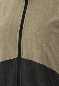 Veste zippée présentant une partie supérieure texturée beige et une partie inférieure noire contrastante, avec une fermeture éclair noire et un tissu ribbed.