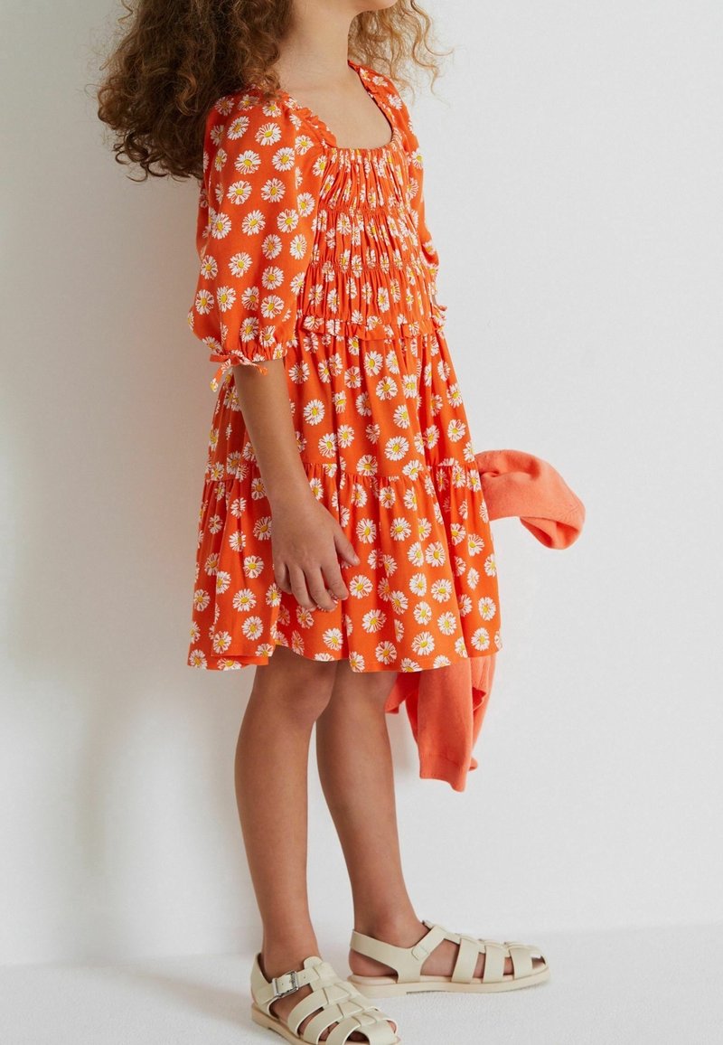 Enfant en robe orange à motifs floraux tenant un pull assorti, portant des sandales blanches fermées, debout devant un fond clair uni.