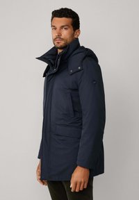 Mężczyzna w granatowym zimowym płaszczu z wysokim kołnierzem i kapturem, stojący bokiem na jednolitym jasnym tle.