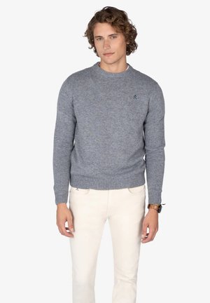 Suéter gris de punto con cuello redondo y puños acanalados. Presenta un pequeño logo bordado en el pecho. Se lleva con pantalones de color claro.