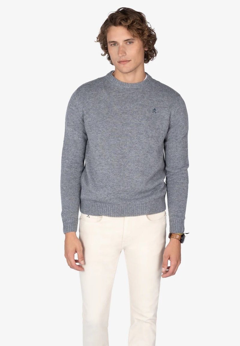 Suéter gris de punto con cuello redondo y puños acanalados. Presenta un pequeño logo bordado en el pecho. Se lleva con pantalones de color claro.