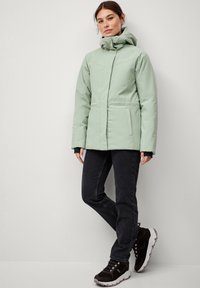 Lys grøn polstret jakke med høj krave, lynlåslukning og to sidelommer, parret med sorte jeans og sorte sneakers med pink accenter.