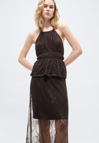 Naine, kes kannab varrukateta tekstureeritud tumedat pruuni kaelaümbrise ja sobiva pitsseeliku läbipaistva pealiskihiga, seisab käed selja taga.