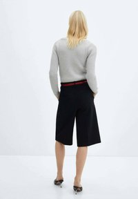 Šedý žebrovaný svetr a černé přiléhavé bermudy s červeným akcentem na pásku. Modelka má na sobě černé boty na podpatku, viděná zezadu.