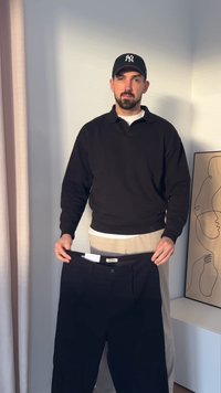 Μαύρο παντελόνι που κρατά ένα άτομο με μαύρο μακρυμάνικο μπλουζάκι και καπέλο. Το παντελόνι διαθέτει κλείσιμο με κουμπί και ετικέτα.