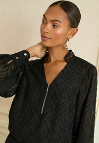 Bluză neagră cu un model texturat ondulat, fermoar în față și mâneci bufante. Prezintă un decolteu înalt cu detalii din dantelă și accente cu nasturi.