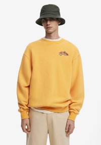 Gul sweatshirt med ribbede manchetter og kant, prydet med et farverigt broderet design på venstre bryst. Modellen bærer en grøn bøttehat.