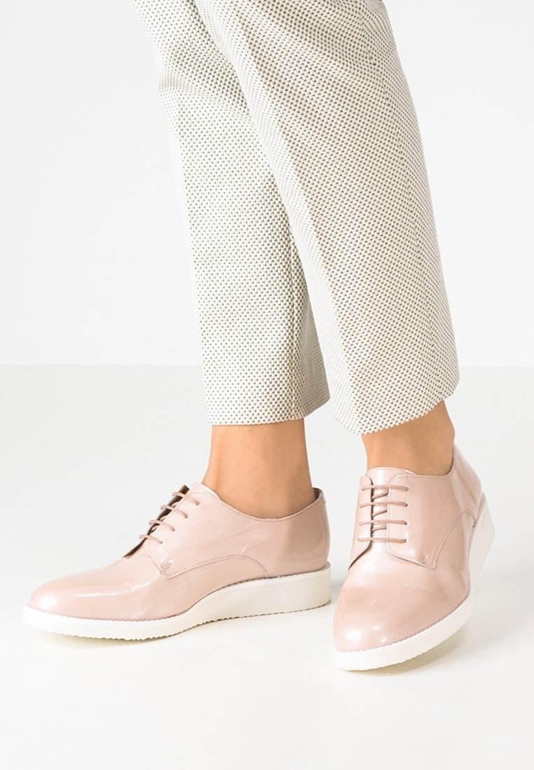 Femme portant des chaussures à lacets en cuir verni rose clair avec des semelles blanches épaisses et un pantalon blanc court à petit motif de points noirs.