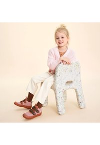 Zapatos de textil rosa con correas y suelas marrones. El niño está sentado en una silla ligera con manchas, vistiendo un suéter de punto rosa y pantalones crema.