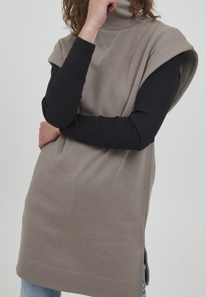 Person iført en ærmeløs beige tunika over en sort ribbet langærmet top, med den ene arm krydset og den anden hånd, der rører ved deres ansigt.
