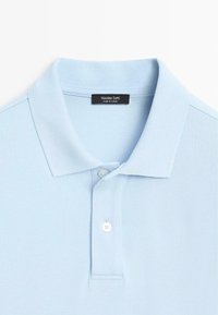 Polo bleu clair à manches courtes avec un col plié, deux boutons blancs et une étiquette noire "Massimo Dutti Made in Turkey" à l'intérieur de l'encolure.