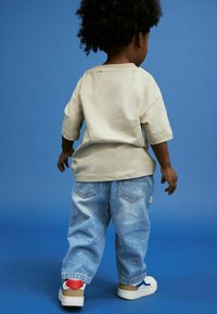 Camicia beige a maniche corte, jeans di denim blu chiaro e sneaker bianche con dettagli rossi e marroni su uno sfondo blu.