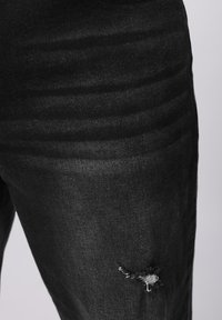 Juodos spalvos džinsai iš džinsinio audinio, turintys pažeistą kelio vietą su plyšiu, lygios tekstūros ir aptemo dizaino. Subtilios išblukusios juostos palei audinį.