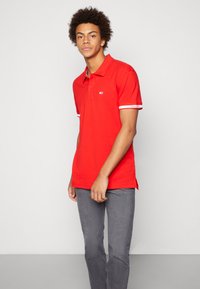 Red polo shirt with a collared neckline, short sleeves featuring white accents, and side slits. Paired with grey pants.