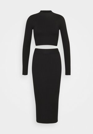 Ensemble deux pièces en maille côtelée noire avec crop top à manches longues et jupe midi taille haute et ajustée sur fond uni.