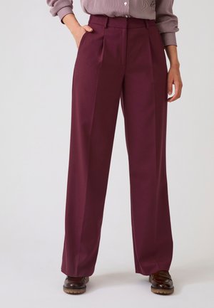 Persoon draagt hoog getailleerde, wijd uitlopende bordeauxrode broek gecombineerd met bruine schoenen en een gestreept overhemd met knopen, hand in de zak.