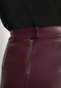 Detailní záběr osoby v černém topu a v bordó kožené sukni s vysokým pasem, na které je viditelný vertikální zip po boku.