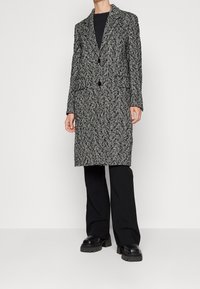 Person, der einen langen Mantel mit Fischgrätmuster in Schwarz und Weiß, ein schwarzes Oberteil, schwarze Hosen und klobige schwarze Plateauschuhe trägt.