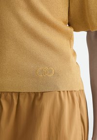 Maglia gialla in maglia con finitura testurizzata; abbinata a una parte inferiore in tessuto liscio color beige. Dettaglio del logo ricamato vicino all'orlo.
