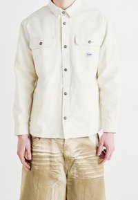 Ljus beige skjorta med knappar, tillverkad av bomull, med två bröstfickor och metallknappar. Bärs över texturerade, ljusbruna byxor.