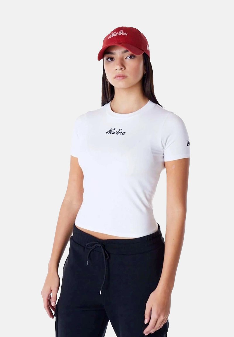 Witte fitted t-shirt met zwart "New Era" logo, gecombineerd met zwarte sweatpants; model draagt een rode baseballpet met logo.