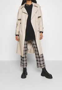 Abrigo trench beige con diseño de doble botonadura, pantalones anchos de cuadros negros y botas negras con cordones. Tela suave y detalles con botones.