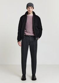 Giacca bomber nera con texture, maglione a coste viola chiaro, pantaloni scuri con motivo, berretto in maglia grigio e stivali in pelle nera.