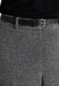I pantaloni grigi in tweed presentano un tessuto testurizzato con motivi sottili. Sono accompagnati da una cintura in pelle nera con una fibbia argentata.