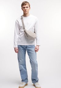 Borsa a tracolla in tessuto beige e morbido indossata a traverso. Abbinata a una camicia bianca a maniche lunghe, jeans azzurri chiari e sneaker color crema. Design semplice.