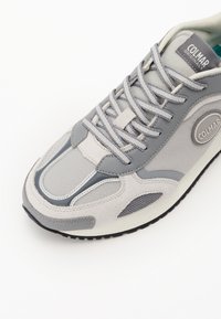 Scarpa sportiva grigia realizzata in materiali mesh e suede. Presenta lacci, un logo rotondo e una suola texture. Design minimalista con accenti a contrasto.