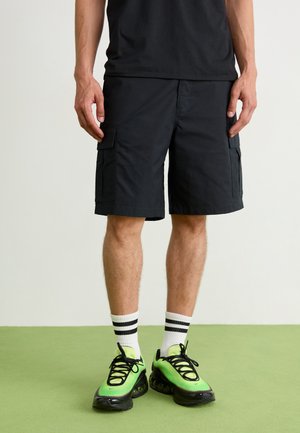 Black cargo shorts made of cotton fabric feature two side pockets. Paired with bright green sneakers and white crew socks with black stripes.
