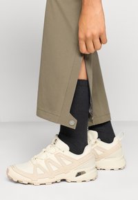 Μπεζ αθλητικά παπούτσια με υφασμάτινη υφή και λείες λεπτομέρειες, συνδυασμένα με σκούρες γκρι κάλτσες και ανοιχτό πράσινο παντελόνι με φερμουάρ.