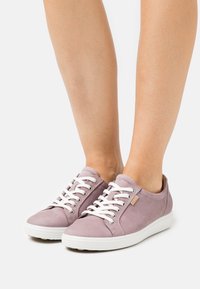 Zapatillas de ante de color morado claro con cordones blancos, suela de goma y parche de logo en color marrón; diseño de corte bajo con puntera redondeada y textura suave.