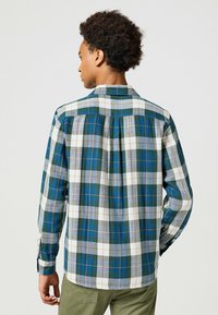 Camisa de franela a cuadros con patrones en verde, blanco y gris, con acentos en naranja, cuello con solapa y puños abotonados.