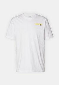 Bílý bavlněný tričko s kulatým výstřihem, krátkými rukávy a žlutým logem "Lacoste" s zeleným krokodýlem na hrudi.