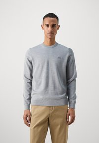 Pull gris en matière tricotée douce, avec un col rond classique, des poignets côtelés et un discret logo sur la poitrine.