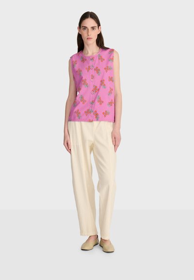 Femme portant un haut sans manches rose à boutons avec imprimé floral orange, pantalon large crème, et chaussures beige à enfiler, debout devant un fond blanc.