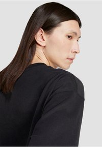 Črna majica s kapuco s krožnim izrezom, mehka tkanina in vidni šivi. Model ima ravne lase in je v profilni poziciji.