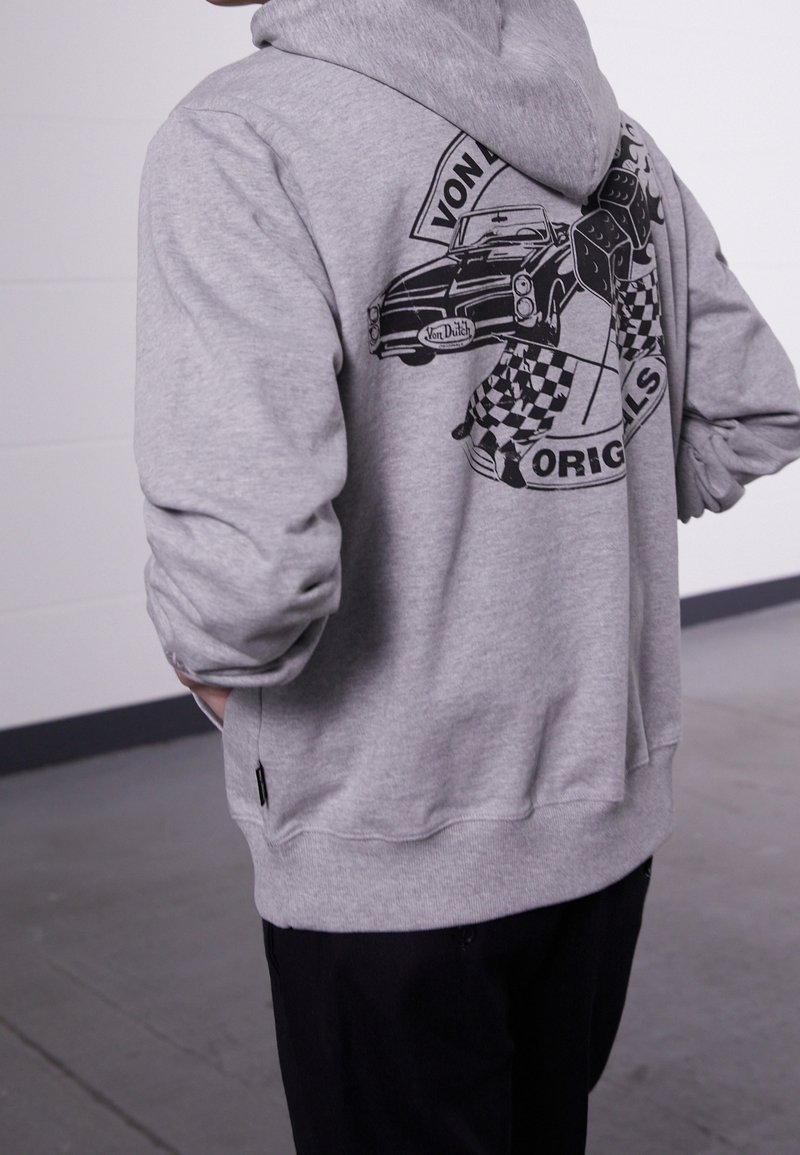 Personne portant un sweat à capuche gris featuring un graphisme noir d'une voiture vintage et des drapeaux à damier avec le texte « Von Dutch Originals » au dos.